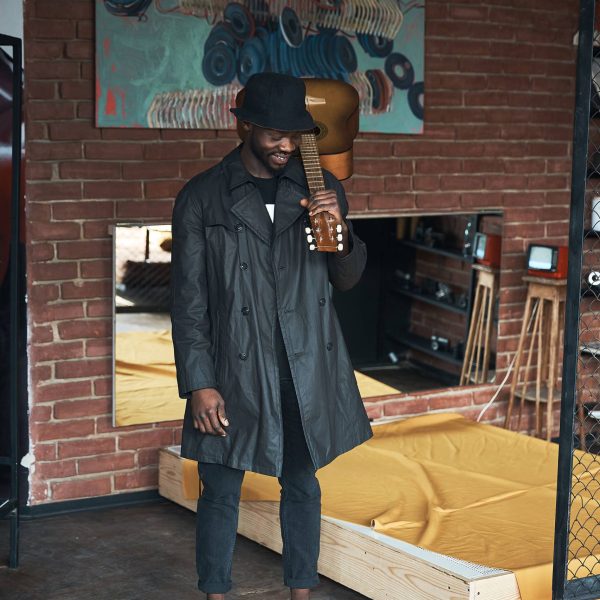 Musician in black trench coat and bucket standing with guitar. Songwriter in loft room. Fashion 2019