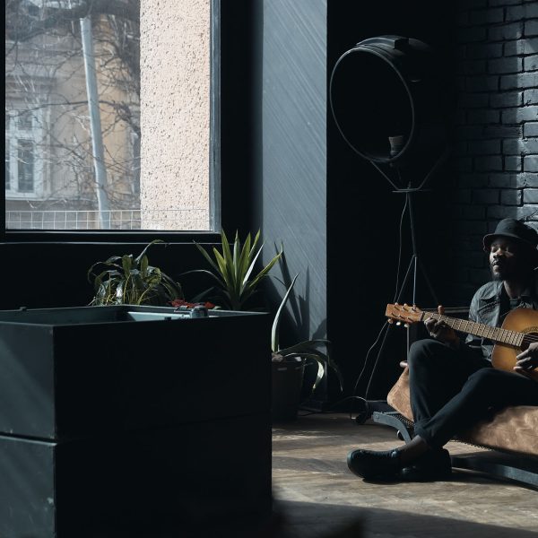 Musician in black trench coat and bucket hat sitting on bed and playing guitar. Songwriter in loft room. Fashion 2019